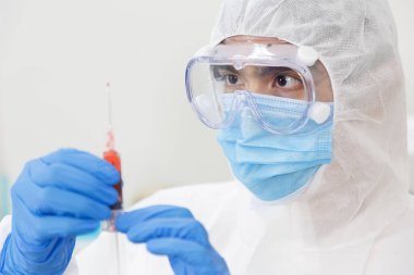 Koruyucu kıyafetli (PPE Suit) ve maskeli Asyalı bir adam şırınga ve aşı taşıyor. Grip aşısı, koronavirüs, ebola, tüberküloz. Biyolojik tehlike. Covid-19 salgını..