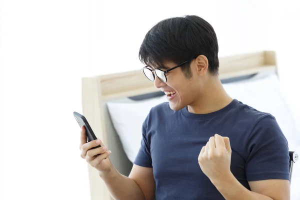 Masada cep telefonu kullanan mutlu Asyalı adam internette iyi haberler okuyor, Asyalı adam heyecanlanıyor zafer çığlığını sevinçle evde zaferi kutluyor. Ev Konseptinden Çalışma. 