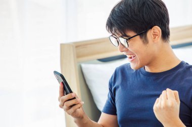 Masada cep telefonu kullanan mutlu Asyalı adam internette iyi haberler okuyor, Asyalı adam heyecanlanıyor zafer çığlığını sevinçle evde zaferi kutluyor. Ev kavramından çalış. Uzayı Kopyala.