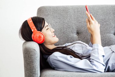 Neşeli Asyalı, kablosuz kulaklık takan akıllı telefondan müzik dinleyen neşeli kadının yan görüntüsü kanepeye uzandı..