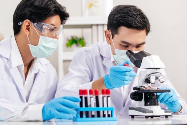 İki erkek bilim adamı yüz maskesi takıyor. Çözümü mikroskop ile kontrol ederken laboratuvarda çalışıyor.