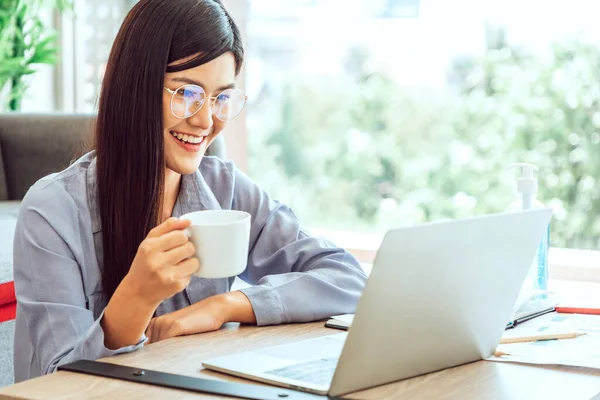 Young Cute Woman feels happy holding a cup of coffee while video call Meeting on Work For Home Period. COVID-19 Lock Down.  Copy Space and Warm Tone.
