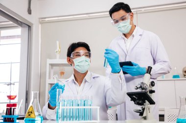 İki erkek bilim adamı yüz maskesi takıyor. Çözümü mikroskop ile kontrol ederken laboratuvarda çalışıyor.