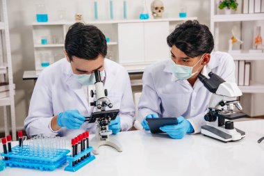 İki erkek bilim adamı yüz maskesi takıyor. Çözümü mikroskop ile kontrol ederken laboratuvarda çalışıyor.