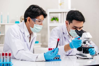 İki erkek bilim adamı yüz maskesi takıyor. Çözümü mikroskop ile kontrol ederken laboratuvarda çalışıyor.