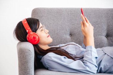 Neşeli Asyalı, kablosuz kulaklık takan akıllı telefondan müzik dinleyen neşeli kadının yan görüntüsü kanepeye uzandı..
