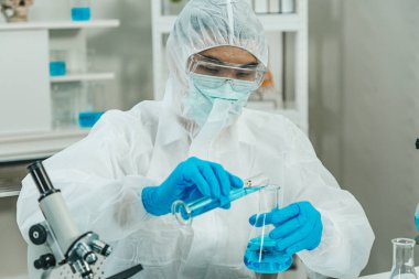 PPE Suit 'te çalışan erkek bilim adamı silindirden Erlenmeyer Flask' e Biyolojik Tehlike İşaretli Çözüm dökerken laboratuvarda çalışıyor. Mavi Ton. SARS-CoV-2, Covid-19 Teması.