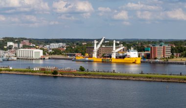 Kiel, Almanya-09.08.2025: Parlak sarı BigLift kargo gemisi Happy Sky rıhtıma yanaştı, vinçler yüklendi, şehir binaları ve yeşil park yaklaştı.