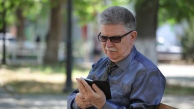 Yaşlı adam belgili tanımlık telefon perde dokunur ve yazın güneşli bir yaz aylarında muhteşem bir sokakta oturan yaptığı telefon internet'te sörf bir kravat mustashe gri saçlı bir adam portresi güzel bir arka sokakta bilgi istiyor