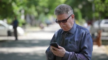 Komuta sizde cep telefonunu görünüyor ve kendine güvenen bir portresi güneşli bir yaz aylarında yeşil bir arka sokakta onun smartphone üzerinde net sörf bir kravat mustashe adamla yaşlı yaz aylarında bir arka sokakta ekran dokunur