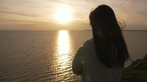 可爱的女孩穿着白色西装看着在夏季的黑海日落后 一个漂亮的女孩与长松散的头发在黑色的海岸边和看金色日落路径在夏季 — 图库视频影像