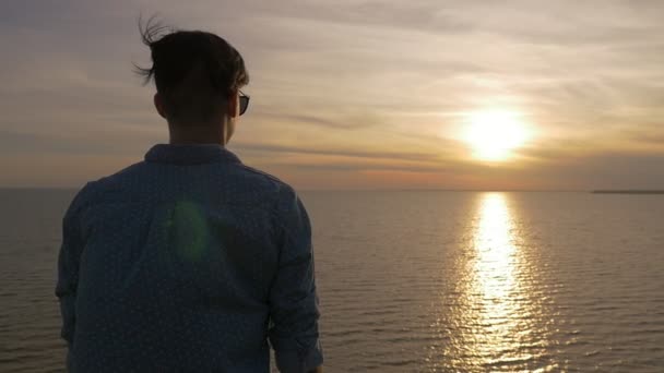 芸術の人が立ちサングラス立ち 夏には美しい夕日を黒海海岸に輝く太陽のパスを見て興奮して若い男の黒海の海岸をバックにきらめく夕日を楽しんでいます — ストック動画