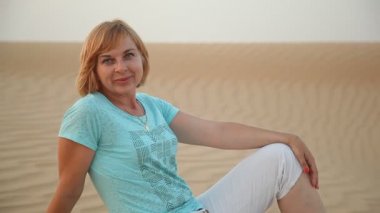 Güneşli bir yaz tişört ve pantolon tatlı dunes Dubai ile yaz aylarında kapalı deniz kıyısında oturan mutlu sarışın bir kadın portresi Dubai kumlu bir sahil neşeli kadın oturur. O iyimser gülümsüyor
