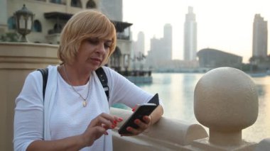 Bir su kanalı ayakta ve telefonunu net sörf bir backpacker sarışın kadın portresi, onun fotoğrafları-Dubai ile şık sarışın kadın görünüyor. O bir su kanalı çit ile ısıtmak, yaz aylarında standları