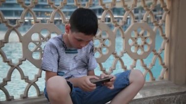 Zeki çocuk yüzme havuzunda oturuyor ve bir video oyunu cep video oyunları oynarken onun smartphon 10 yaşında bir çocuk özgün bir bakış oynar. O bir T-shirt olmak ahşap bir çit oturur ve yaz aylarında şort