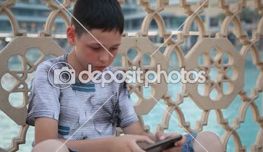 Küçük çocuk bir yüzme havuzunda oturuyor ve bir video oyunu onun smartphone vasıl vasıl onun hareket eden video oyunları oynarken 10 yaşında bir çocuk heyecan verici bir görünümünü oynar. O bir T-shirt giymiş figüratif bir çit oturur ve yaz aylarında şort