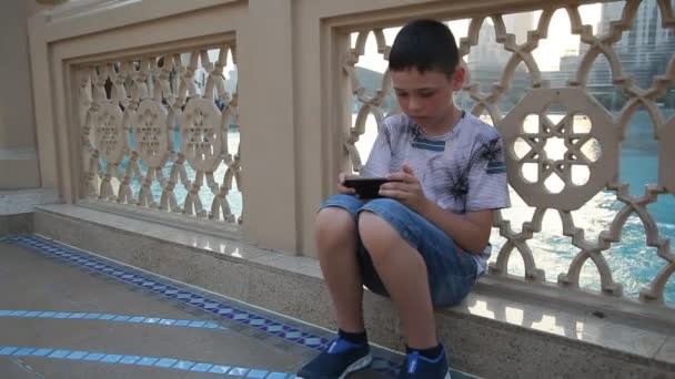 一个活泼的男孩坐在路堤上 在迪拜玩游戏视频一个10岁的男孩在夏天坐在迪拜的一个水上通道码头上 在他的智能手机虚拟现实中暴跌的情绪视图 — 图库视频影像