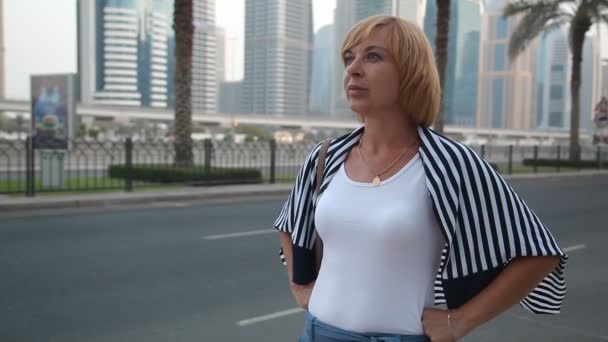 时尚的女性游客等待在高速公路上的人在迪拜的摩天大楼 一个漂亮的女人穿着白色 牛仔裤和条纹斗篷等待一个阳光明媚的日子在迪拜高速公路上的某人令人兴奋的看法 — 图库视频影像