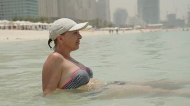Rahatlatıcı kadın mavi deniz sularında Dubai yaz yavaş hareket profil bir beyaz kap ve deniz suları kıyısında oturan ve Dubai'deki gökdelenler slo-mo ile dalgalar deniz bakarak bikini içinde mutlu bir kadın oturuyor