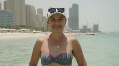 Neşeli kadın bikini deniz plaj Dubai ve yaz beyaz bir şapka ve güneş gözlüğü ve kumlu bir plaj üzerinde duran ve sonsuza dek mutlu yaz aylarında Dubai'de gülümseyen bikini neşeli ve şık bir kadın portresi gülümser durumda