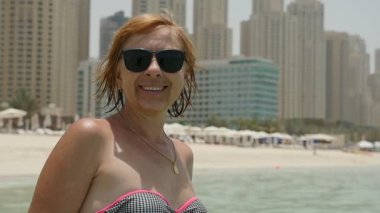 Neşeli kadın bikini deniz plaj Dubai ve yavaş hareket güneş gözlüğü ve kumlu bir plaj üzerinde duran ve Dubai'de gökdelenler içinde belgili tanımlık geçmiş ile yaz aylarında gülümseyen bikini mutlu sarışın bir kadın portresi gülümser durumda