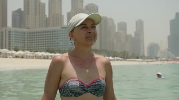 比基尼微笑的女人站在迪拜的海边与建筑物在夏季的照片一个开朗的女人比基尼和一个白色的帽子站在沙滩上的蓝色海水和微笑在夏季在迪拜慢动作 — 图库视频影像