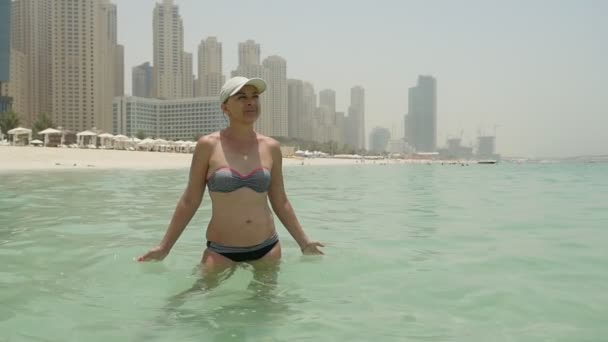 穿着比基尼的休闲女郎站在迪拜的海边 夏天的摩天大楼上画着一个穿着比基尼的乐观女人 戴着白色的帽子和太阳镜 拍着海水 在迪拜的沙滩上站着 — 图库视频影像