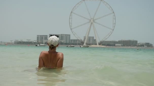 放松的女人坐在蓝色的海水中 看着在迪拜的摩天轮在斯洛伐克 莫一个时髦的女人坐在海滩上的海水 并观察在夏季在迪拜的高摩天轮慢动作的一个时尚女性的后视图 — 图库视频影像