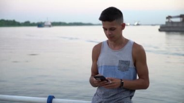 Genç adam standları ve ayakta ve günbatımında bir setin küpeşte ile onun smartphone ekran dokunaklı bir singlet içinde mürettebat saç modeli ile yaz genç bir adam portresi batımında telefonunu bir nehir quay adlı dokunur 