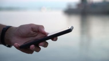 Adamın elini yaz portre siyah telefon tutmak ve güzel günbatımı, bir nehir iskele üzerinde bazı bilgi arayan onun ekran dokunaklı bir erkek el günbatımında bir smartphone nehir üzerinde net tarama