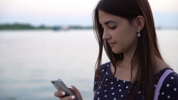 美丽的女孩站在她的手机上 在一个河码头上发短信一个漂亮的女孩与长头发冲浪网智能手机在河岸上的一条漂浮的船在日落 — 图库视频影像