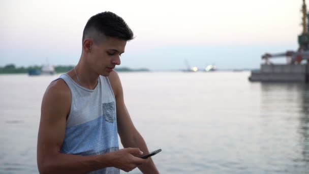 若い男が立ち 立っていると素敵な夕日に船の絵のような堤防に彼のスマート フォンでネットをブラウズして白一重項の陽気な男のプロファイルの夏の夕日の河岸で携帯電話の画面に触れる — ストック動画