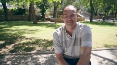 Bir bankta oturan ve mutlu yaz bir bankta oturan ve heyecanla yeşil ağaçlar ve güneşli çimleri park sokakta gülümseyen gözlük Avrupa'dan modern bir adam portresi yeşil bir parkta gülümseyen neşeli adam