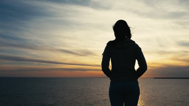 梦想中的女孩看着黑色的海水与灿烂的太阳路径返回一个温柔的年轻女子在一个柔和的运动西装站在一个绿色的海滨 并期待在夏季的阳光路径的神奇日落 — 图库视频影像