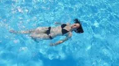 Neşeli bir genç kadının slo-mo bir yüzme havuzunda bir havuzda bir kuş bakışı bir gülümseyen ince kadın mavi mavi sularda yalan ve yavaş hareket bir havuzunda eğlenirken siyah bikini yalan hava atış 