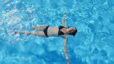 Mutlu genç kadın swinning hava atış bir kuş bakışı gök mavi sularda yalan ve yavaş bir havuzda eğlenceli siyah bikini neşeli bir genç kadının slo-mo bir havuzda sırtüstü 