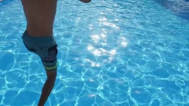 陽気な少年ジャンプ フィート最初スローモーション スローモーションで夏には広々 としたプールのシアン ブルー水にジャンプの長いショート パンツでティーンエイ ジャーの陽気なビューで空の青色の水でプールで — ストック動画