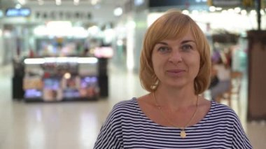 Gülümseyen ve büyük bir salonda vitrinler çizgili bir bluz neşeyle gülümseyen ve insanlarla bir cam gibi eğlence merkezinde duran bir kendinden emin ve iyimser sarışın kadın portresi ile ayakta neşeli sarışın kadın 