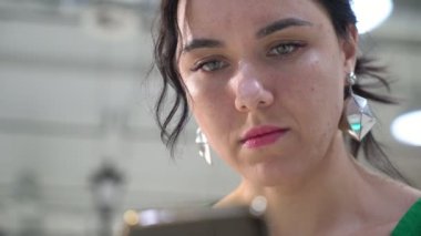 Çekici esmer kadın net bir salonda oturmuş ve onun smartphone üzerinde net sörf şık küpe ile gülümsemek gülümseyen genç bir kadının uzun ve dalgalı saçlı bir salonda oturan telefonunda tarama