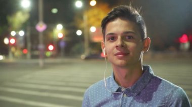 Neşeli adam gece bir teşvik sportif brunet adam portresi, bir zebra geçiş müzik ayakta sonbaharda bulanık ışıklı bir şehirde kulaklık aracılığıyla müzik dinlemek bir mürettebat saç kesimi ile dinliyor