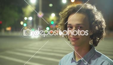 Bir zebra geçiş ayakta ve bir şehirde bir zebra geçiş trafik ile ayakta ve mutlu bir şehirde sokak sonbahar gece gülümseyen uzun dalgalı saçlı Genç bir adam portresi geceleri gülümseyen glamour sarışın adam  