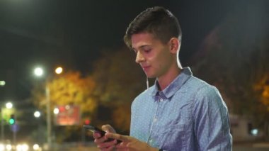 Sportif adam net sörf ve gece romantik bir brunet adam müzik ve onun akıllı telefon net sonbahar gece kızına bazı metin gönderirken tarama profili lampposts ayakta