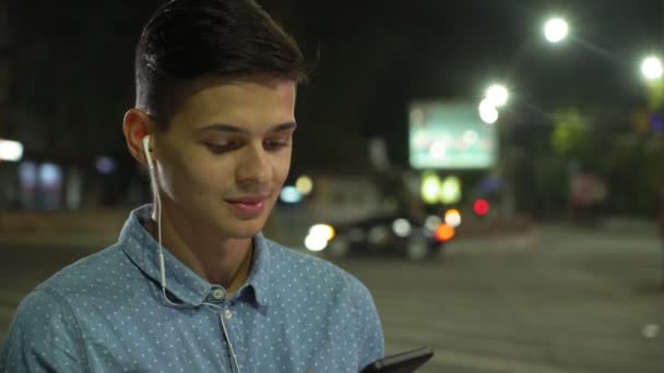 整洁的年轻人正在网上冲浪 晚上站在灯柱前的一个运动的黑发男人在他的智能手机上听音乐和浏览网的简介 同时在秋天的晚上给他的爱人发一些短信 — 图库视频影像