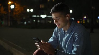 Genç adam şehir kaldırım kaldırım kenarında oturuyor ve gece heyecanlı görünümü onun akıllı telefon ekranına bakarak ve biraz bilgi arayan neşeli bir brunet insanın net gözatabilir. O müziği dinliyor ve geceleri çok güzel  