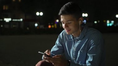Brunet delikanlı müzik ve onun telefon oturma ve sonbahar gece sokağın ortasında gülümseyen sportif genç bir adamın gece etkileyici manzaraya, internette sörf. O smartphone net taradığında ve kulaklıkları.