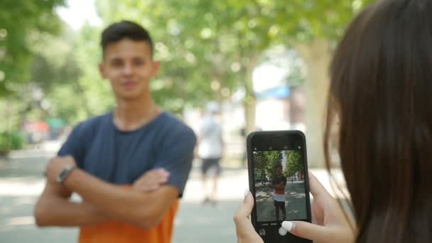 漂亮的女孩拍摄她的愉快的男朋友与她的手机谁在斯洛伐克 莫的原始视图的微笑深色与交叉武器在夏天的公园里 他的宠儿在斯洛伐克 他的形象在银幕上可见 — 图库视频影像