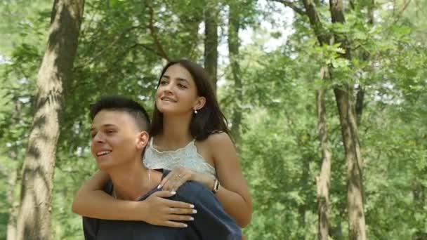 幸せな若い男は 夏緑の森で楽しい若いカップルの陽気な眺めで緑の森で彼の背中に彼のガール フレンドを運びます 男らしい男は彼の背中に彼の笑顔の姫を保持し 幸せを感じています — ストック動画