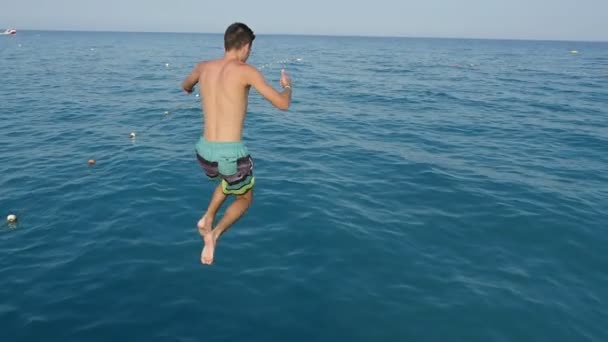 活跃的黑发男子在夏天在海上首先跳跃脚在慢动作令人兴奋的看法一个运动的黑发男子穿着彩色短裤跳脚首先在蓝色的海浪从码头在夏天的一个码头在慢动作 — 图库视频影像