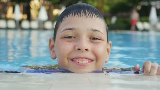 スローモーション スローモーションで夏にターコイズ ブルーの海と周りの木々 の泳ぐ池で喜んで口を開けて笑っている陽気な少年のポートレートで夏のプールの端に幸せな少年の笑顔 — ストック動画