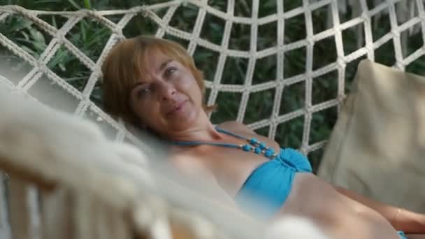 Femme souriante se balançant dans un hamac exotique dans les ombres des arbres à Pékin Vue joyeuse d'une femme blonde heureuse dans un bikini bleu relaxant, souriant et couché dans un hamac fait de cordes blanches fortes par une journée ensoleillée à Pékin  